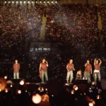 `BTS performing on stage before a packed stadium crowd during their ARIRANG World Tour, with thousands of fan lightsticks glowing in the darkness.`
