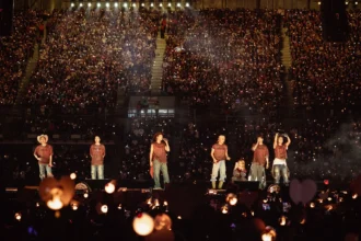 `BTS performing on stage before a packed stadium crowd during their ARIRANG World Tour, with thousands of fan lightsticks glowing in the darkness.`