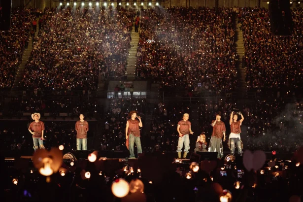 `BTS performing on stage before a packed stadium crowd during their ARIRANG World Tour, with thousands of fan lightsticks glowing in the darkness.`