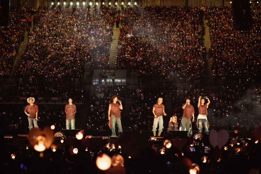 `BTS performing on stage before a packed stadium crowd during their ARIRANG World Tour, with thousands of fan lightsticks glowing in the darkness.`