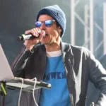Beastie Boys' Mike D performs on stage with a microphone, wearing a blue beanie, sunglasses, and a blue graphic tee under a grey jacket, ahead of his first solo UK show and new single 'What We Got' in 2026