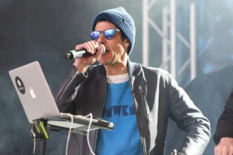 Beastie Boys' Mike D performs on stage with a microphone, wearing a blue beanie, sunglasses, and a blue graphic tee under a grey jacket, ahead of his first solo UK show and new single 'What We Got' in 2026