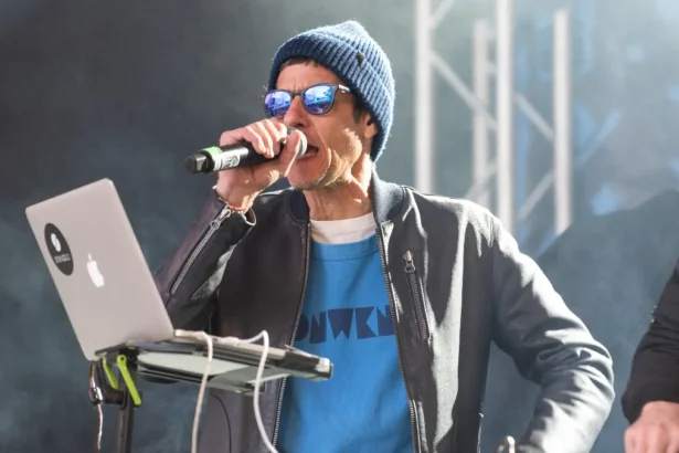 Beastie Boys' Mike D performs on stage with a microphone, wearing a blue beanie, sunglasses, and a blue graphic tee under a grey jacket, ahead of his first solo UK show and new single 'What We Got' in 2026