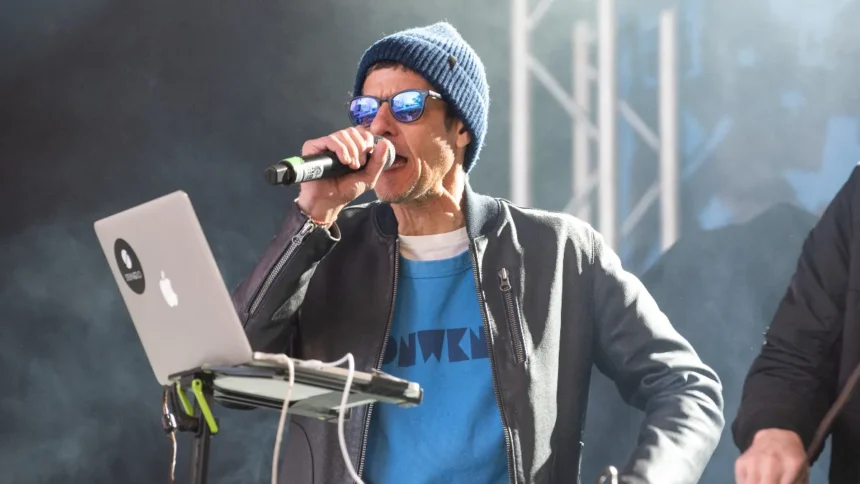 Beastie Boys' Mike D performs on stage with a microphone, wearing a blue beanie, sunglasses, and a blue graphic tee under a grey jacket, ahead of his first solo UK show and new single 'What We Got' in 2026
