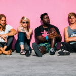 Bloc Party's four members seated casually against a bright pink wall in an outdoor promotional photo shoot