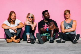 Bloc Party's four members seated casually against a bright pink wall in an outdoor promotional photo shoot