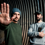 Boards of Canada's Mike Sandison and Marcus Eoin photographed against a corrugated metal tunnel backdrop, Sandison extending his hand toward the camera, 2013 press photo