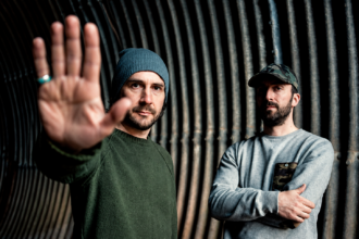 Boards of Canada's Mike Sandison and Marcus Eoin photographed against a corrugated metal tunnel backdrop, Sandison extending his hand toward the camera, 2013 press photo