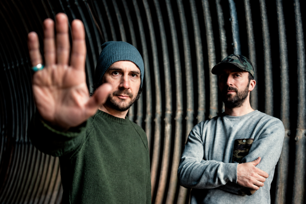 Boards of Canada's Mike Sandison and Marcus Eoin photographed against a corrugated metal tunnel backdrop, Sandison extending his hand toward the camera, 2013 press photo