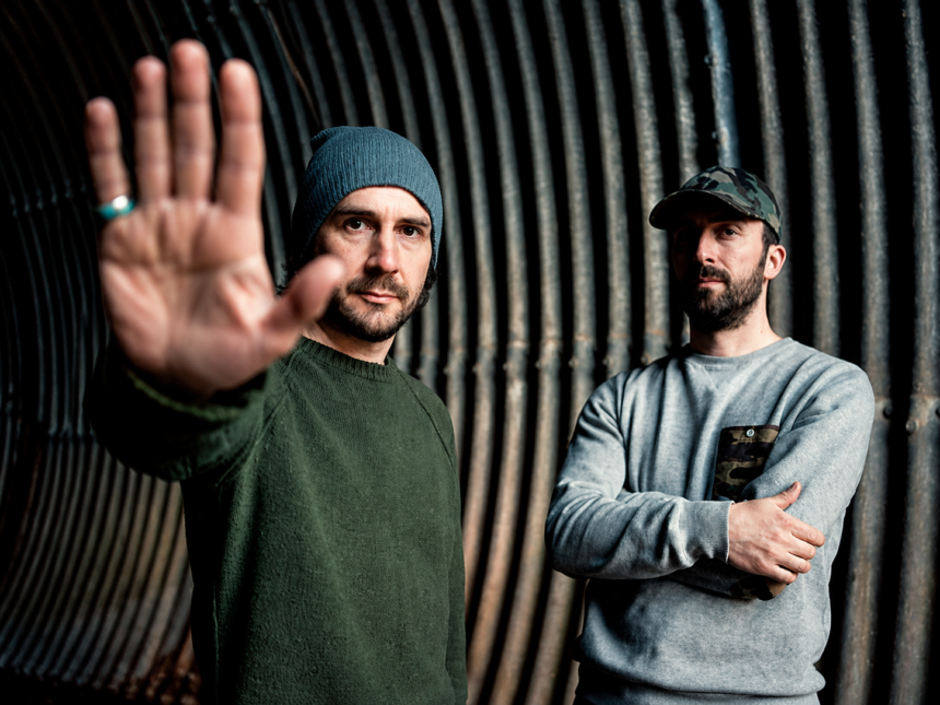 Boards of Canada's Mike Sandison and Marcus Eoin photographed against a corrugated metal tunnel backdrop, Sandison extending his hand toward the camera, 2013 press photo