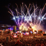 Boomtown Fair 2026 night aerial view showing fireworks, fire effects, and a massive crowd gathered around illuminated stage structures at Matterley Estate, Hampshire, UK