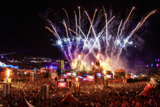 Boomtown Fair 2026 night aerial view showing fireworks, fire effects, and a massive crowd gathered around illuminated stage structures at Matterley Estate, Hampshire, UK