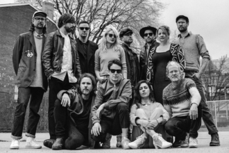 Broken Social Scene band photo, a large group of roughly a dozen musicians posed together outdoors in a parking lot or schoolyard, shot in black and white