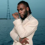 Burna Boy in a cream suit with polka-dot trim, posed with arms crossed against an ocean backdrop