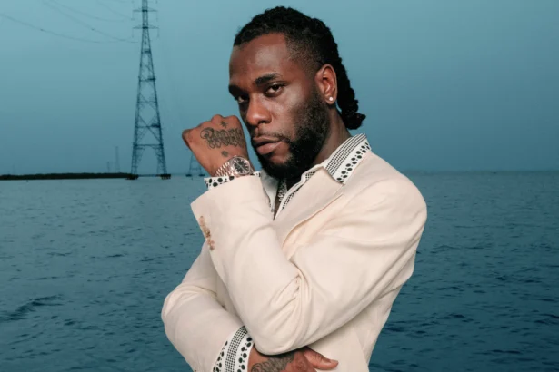 Burna Boy in a cream suit with polka-dot trim, posed with arms crossed against an ocean backdrop