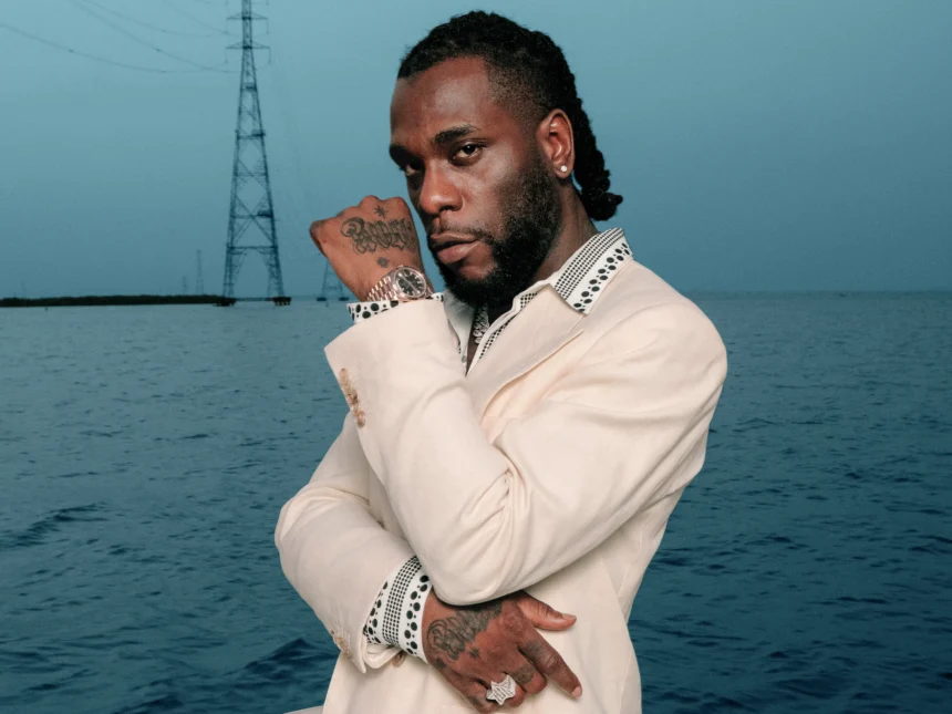 Burna Boy in a cream suit with polka-dot trim, posed with arms crossed against an ocean backdrop