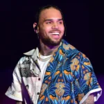 Chris Brown smiling on stage wearing a blue and orange floral open shirt over a white tee and gold chain.