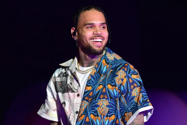 Chris Brown smiling on stage wearing a blue and orange floral open shirt over a white tee and gold chain.