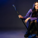 Courtney Hadwin press photo — seated in a purple blazer and platform boots, holding a vintage microphone stand against a moody blue-lit studio background.