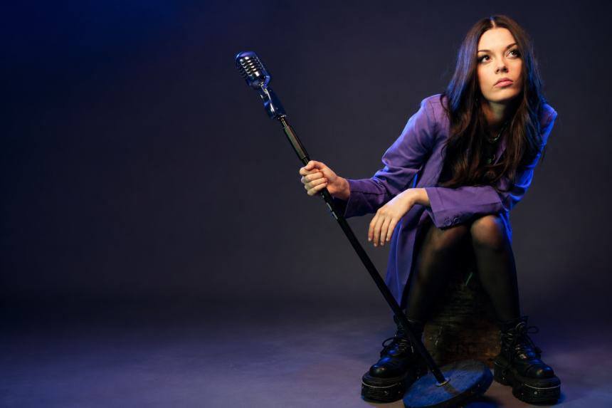 Courtney Hadwin press photo — seated in a purple blazer and platform boots, holding a vintage microphone stand against a moody blue-lit studio background.