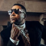 Don Omar poses in reflective sunglasses, a black blazer, and a white turtleneck in front of an ornate stone fireplace, creating a sophisticated and confident portrait.