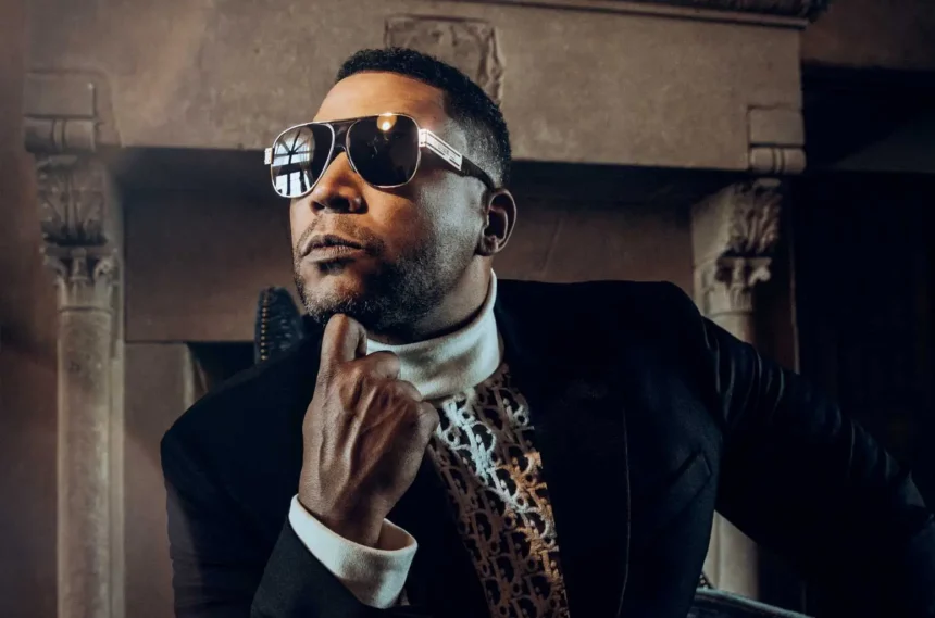 Don Omar poses in reflective sunglasses, a black blazer, and a white turtleneck in front of an ornate stone fireplace, creating a sophisticated and confident portrait.