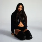 Eartheater sits on a white studio floor with long dark hair, wearing a navy cropped sweater, black shorts, and black over-the-knee leather boots, gazing directly at the camera.