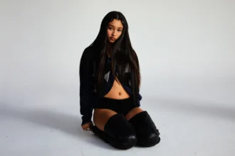 Eartheater sits on a white studio floor with long dark hair, wearing a navy cropped sweater, black shorts, and black over-the-knee leather boots, gazing directly at the camera.