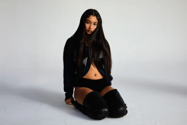 Eartheater sits on a white studio floor with long dark hair, wearing a navy cropped sweater, black shorts, and black over-the-knee leather boots, gazing directly at the camera.