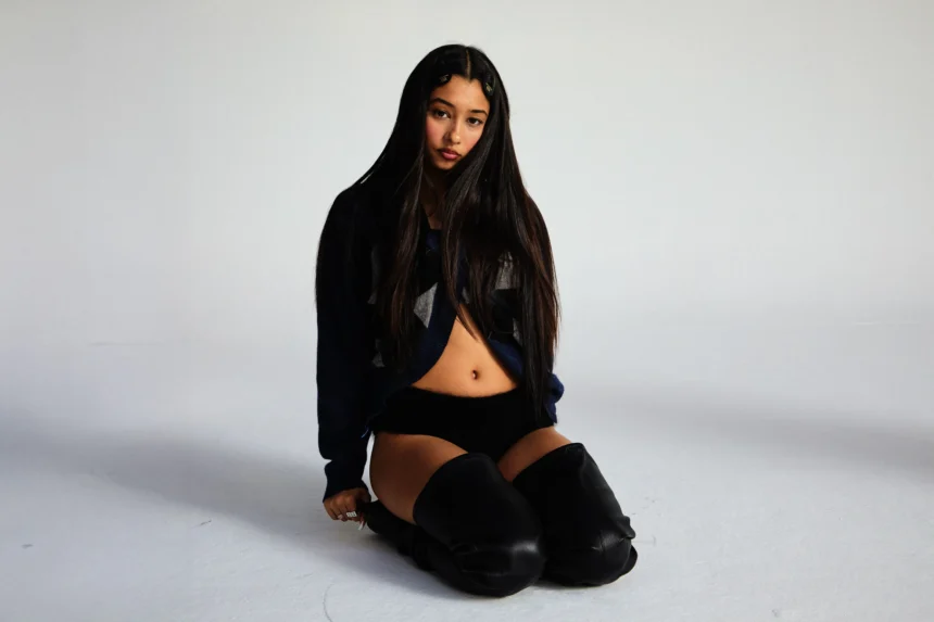 Eartheater sits on a white studio floor with long dark hair, wearing a navy cropped sweater, black shorts, and black over-the-knee leather boots, gazing directly at the camera.