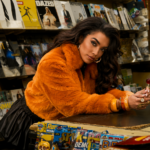 `Erica-Cody leans on a newsstand counter in an orange faux fur jacket holding a lollipop, in a press photo for her new single 'Last Man Standing.'`