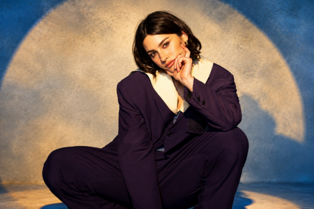 Gracie Abrams poses in an oversized dark purple suit against a soft blue backdrop with a glowing circular light, in a press photo for her 'Look at My Life Tour' and third album 'Daughter From Hell,' out July 17, 2026