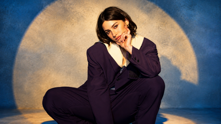 Gracie Abrams poses in an oversized dark purple suit against a soft blue backdrop with a glowing circular light, in a press photo for her 'Look at My Life Tour' and third album 'Daughter From Hell,' out July 17, 2026