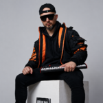 **ALT TEXT:** `HZPROD sits on a white cube holding a mini keyboard synth, wearing black and orange streetwear jacket, black hoodie, snapback cap, and dark sunglasses, with an Urban Barz magazine resting against the cube beneath him`