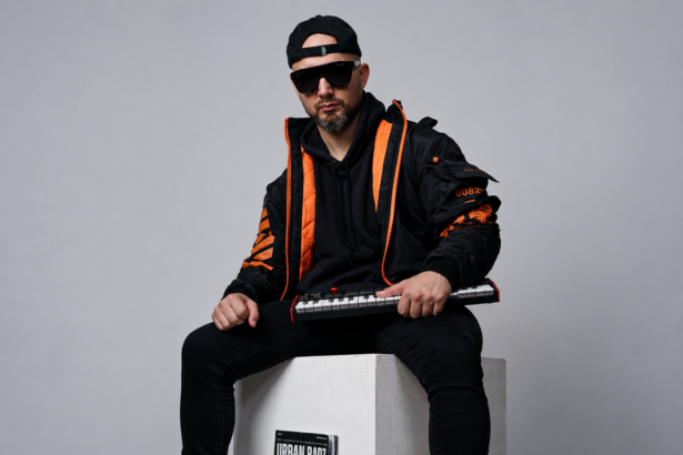 **ALT TEXT:** `HZPROD sits on a white cube holding a mini keyboard synth, wearing black and orange streetwear jacket, black hoodie, snapback cap, and dark sunglasses, with an Urban Barz magazine resting against the cube beneath him`
