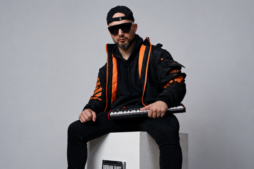 **ALT TEXT:** `HZPROD sits on a white cube holding a mini keyboard synth, wearing black and orange streetwear jacket, black hoodie, snapback cap, and dark sunglasses, with an Urban Barz magazine resting against the cube beneath him`