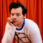 Harry Styles poses against a red curtain backdrop wearing a white fuzzy striped sweater, bold blue beaded necklace, and stacked gold rings, resting his chin on his hand