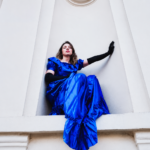 Honddu's Holly Müller perched on a white architectural ledge in a royal blue ruffled gown and long black gloves, shot from below against a pale white building.