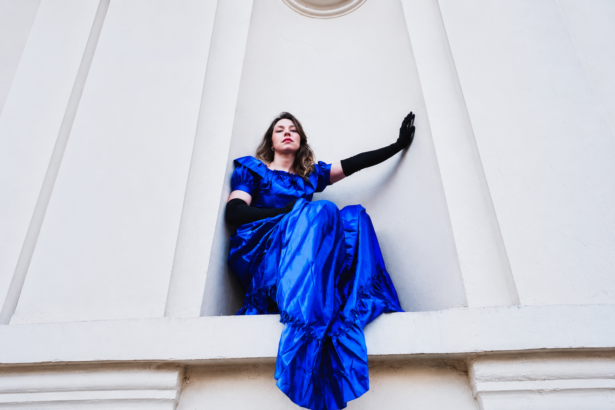 Honddu's Holly Müller perched on a white architectural ledge in a royal blue ruffled gown and long black gloves, shot from below against a pale white building.