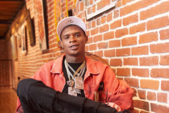 `HoodTrophy Bino rapper wearing orange jacket and gold chains promoting new album Made A Way 2026`