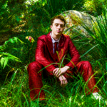 Jackson Mercer sits among lush green grass and foliage wearing a bold red patterned suit and tie, in a press photo accompanying his debut single "Anxiety," released May 27, 2026