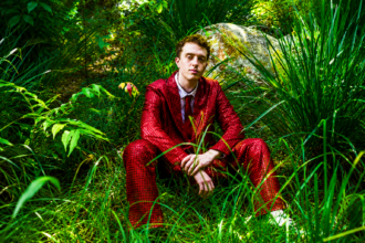 Jackson Mercer sits among lush green grass and foliage wearing a bold red patterned suit and tie, in a press photo accompanying his debut single "Anxiety," released May 27, 2026