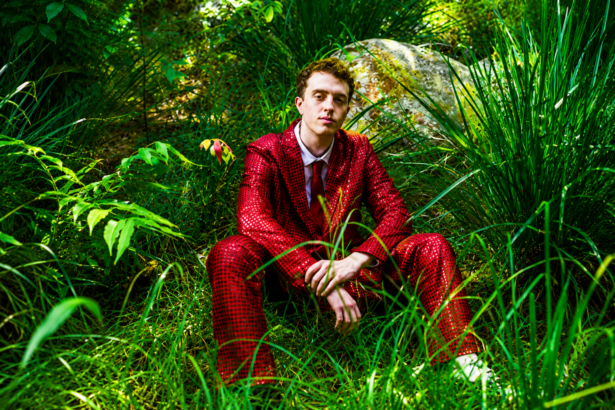 Jackson Mercer sits among lush green grass and foliage wearing a bold red patterned suit and tie, in a press photo accompanying his debut single "Anxiety," released May 27, 2026