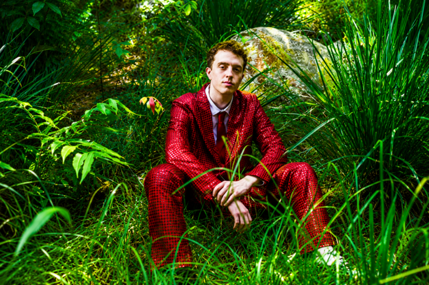 Jackson Mercer sits among lush green grass and foliage wearing a bold red patterned suit and tie, in a press photo accompanying his debut single "Anxiety," released May 27, 2026