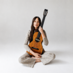 Kacey Musgraves sits cross-legged on a white studio floor, holding a vintage classical guitar upright against her shoulder, wearing a patterned gray outfit.