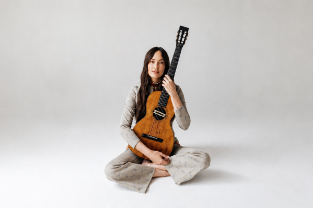 Kacey Musgraves sits cross-legged on a white studio floor, holding a vintage classical guitar upright against her shoulder, wearing a patterned gray outfit.