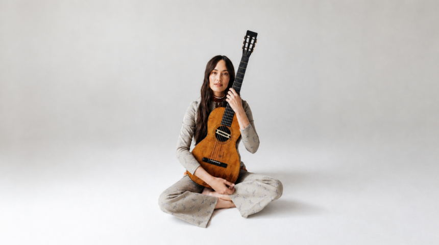 Kacey Musgraves sits cross-legged on a white studio floor, holding a vintage classical guitar upright against her shoulder, wearing a patterned gray outfit.