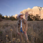 Kalsey Kulyk stands in a grassy desert landscape wearing a tan western outfit and white cowboy hat with mountains glowing in the background for her single “Little Boy.”