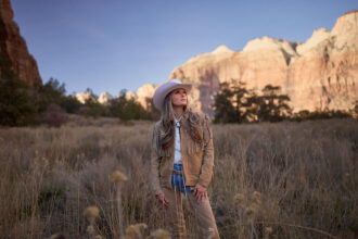 Kalsey Kulyk stands in a grassy desert landscape wearing a tan western outfit and white cowboy hat with mountains glowing in the background for her single “Little Boy.”