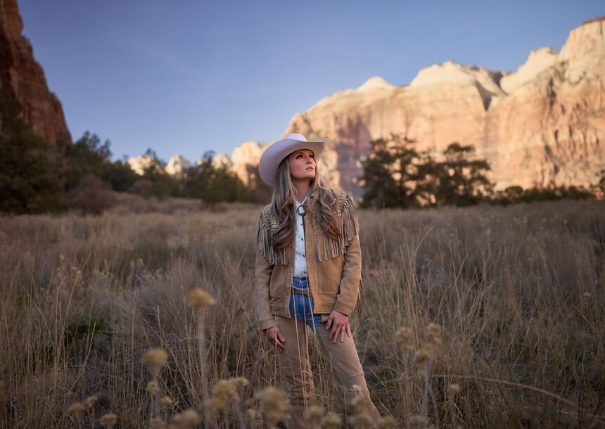 Kalsey Kulyk stands in a grassy desert landscape wearing a tan western outfit and white cowboy hat with mountains glowing in the background for her single “Little Boy.”